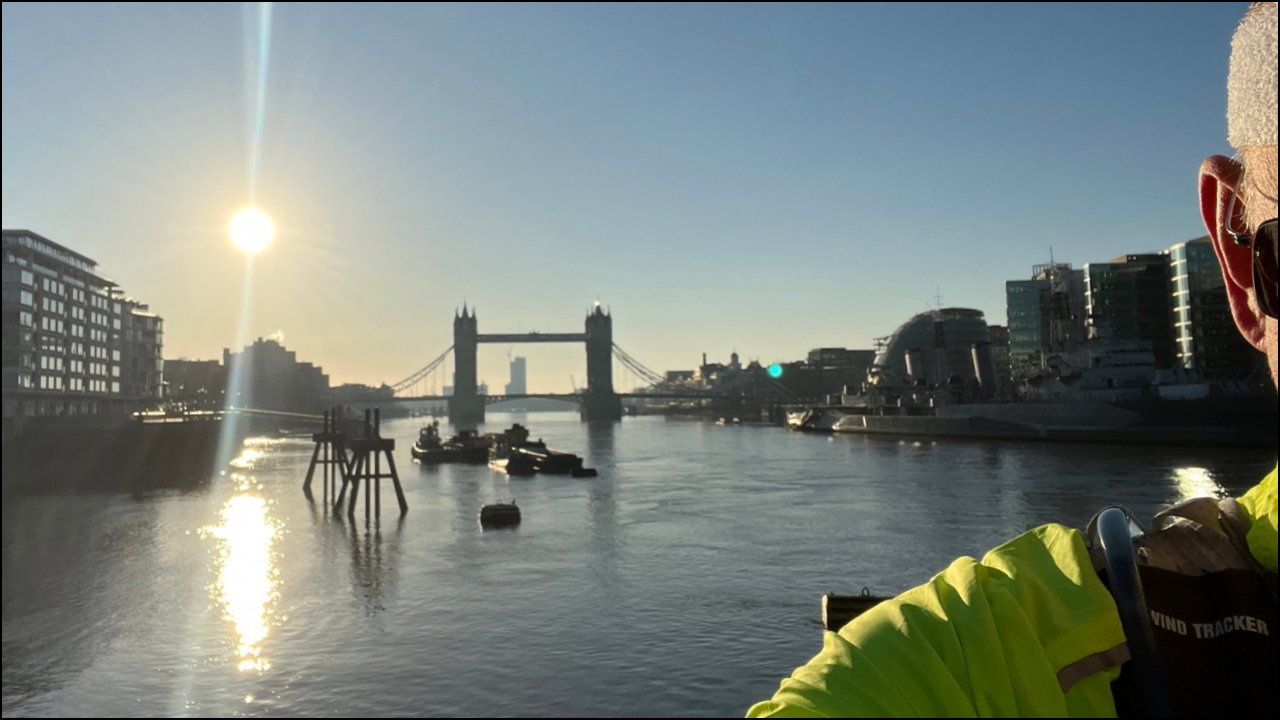 A photograph of the River Thames and Tower Bridge at sunrise with a perfect blue sky. Taken at 7.21am on Sunday 16 March 2025. The Sun is up and dominates the photo just above the buildings to the left of the bridge. Scott Rochester is standing to the right hand side of the photo, deliberately hiding his face. He has a dayglo yellow running top, a runner